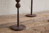 Two Mawindi Candle Holders in rust by nkuku sit on a wooden surface against a beige textured wall, their round bases and lower stems adding charm to earthy decor.