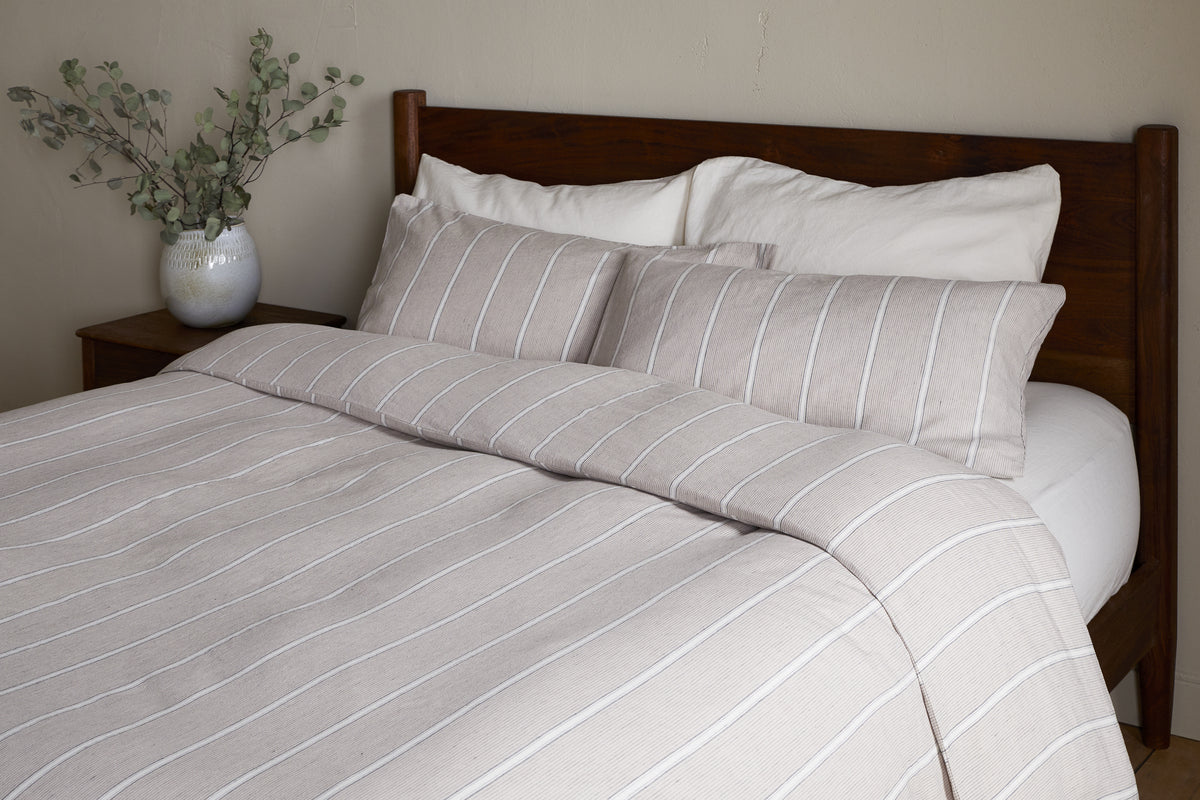 A neatly made bed featuring the nkuku Mora Hemp & Cotton Duvet Cover in Stone Grey, paired with two large pillows and a wooden headboard. A white vase with leafy branches sits on a bedside table against a neutral wall.