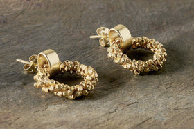 The Mula Earrings - Gold by nkuku are textured, irregular gold-plated hoops with faceted gold beads. These hypoallergenic studs have butterfly backs, a shiny matte finish, and are displayed on a stone surface.