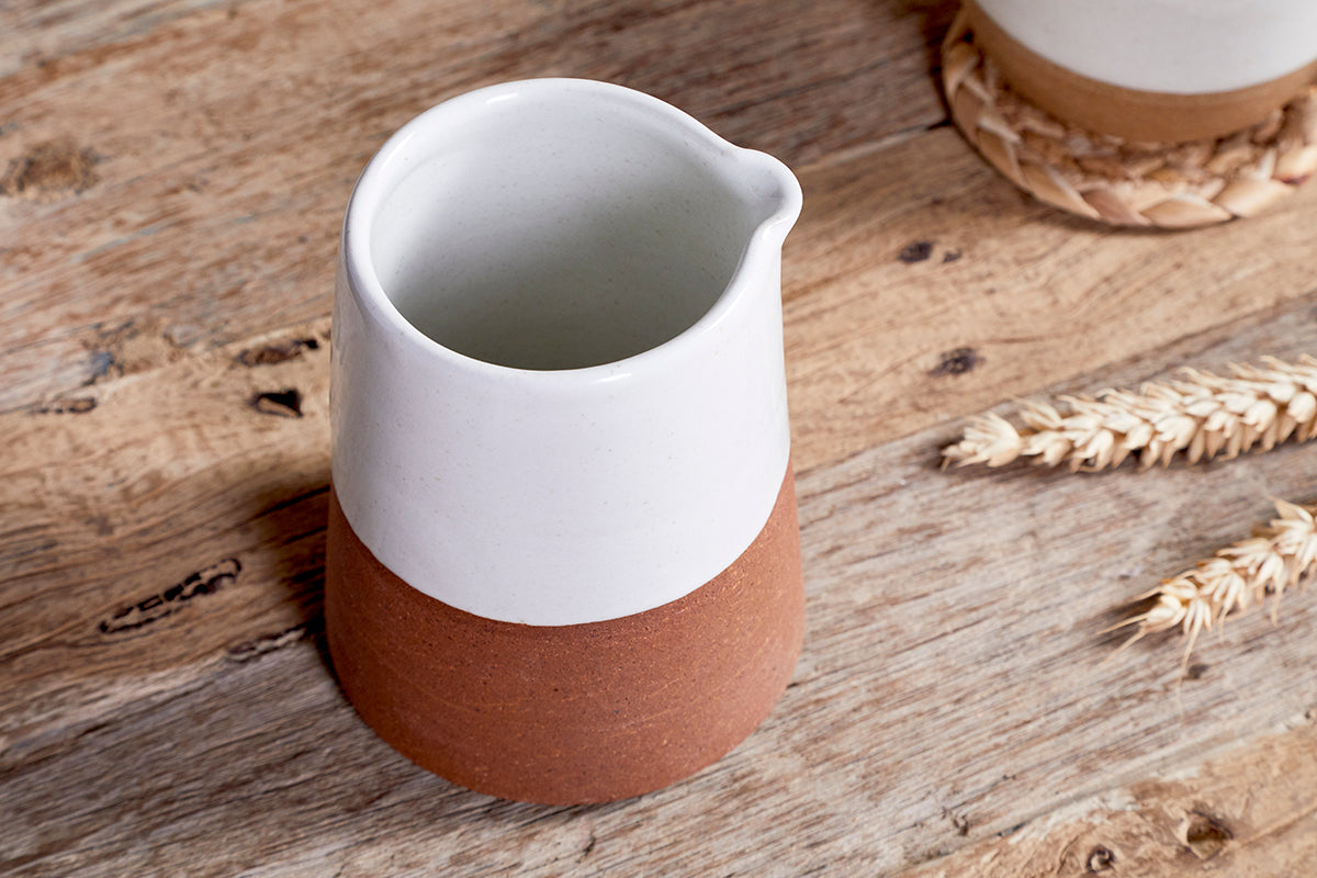 The nkuku Mali Slanting Jug - White, featuring a white top and unglazed brown base, rests on rustic wood with dried wheat and another vessel on a woven coaster, creating a cozy, natural ambiance.