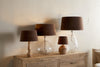 Four Mendai Velvet & Linen Lampshades in Mocha by nkuku are arranged on a wooden sideboard; two have clear glass bases, one has a ribbed ceramic base, and one has a wooden base, all against a light-colored wall.