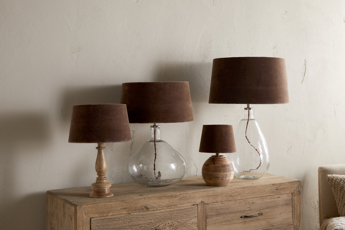 Four nkuku Mendai Velvet & Linen Lampshades in mocha are displayed on a rustic wooden cabinet. Three have transparent glass bases, and one has a textured ceramic base, set against a plain light-colored wall.