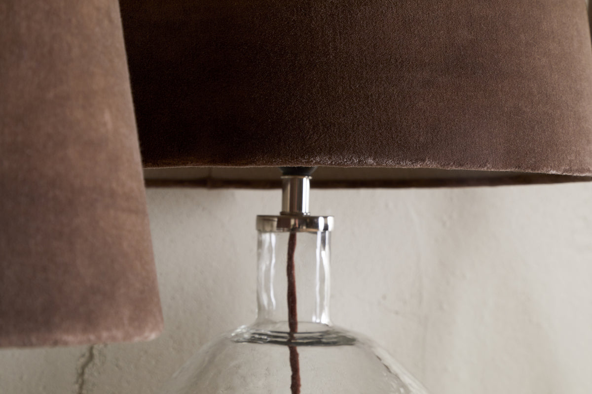 Close-up of the nkuku Mendai Velvet & Linen Lampshade in Mocha, paired with a clear glass lamp and visible cord, set against a textured white wall for a warm, inviting look.