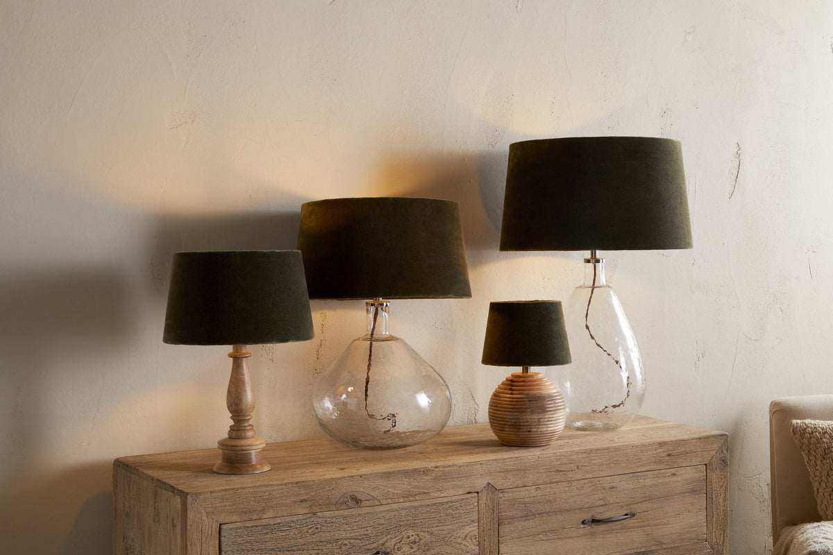 Four Mendai Velvet & Linen Lampshades in Moss Green by nkuku, with glass or ceramic bases, stand on a rustic wooden sideboard, casting soft, warm light and elegant shadows against a textured beige wall.