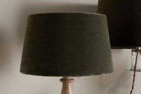 A close-up of the nkuku Mendai Velvet & Linen Lampshade in Moss Green on a wooden base, set against a light wall, with a second similar lampshade partially visible in the background on the right.