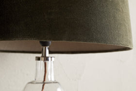 Close-up of the nkuku Mendai Velvet & Linen Lampshade in Moss Green, featuring a textured velvet finish. The glass base and cord are partially visible against a plain, light-toned wall that highlights the lamp's elegant details.