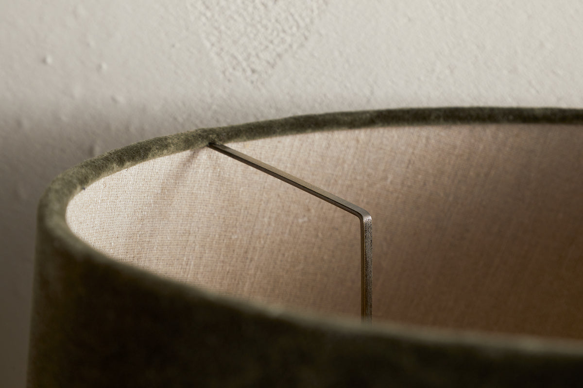 Close-up of the Mendai Velvet & Linen Lampshade by nkuku in Moss Green, highlighting its textured velvet exterior, beige linen interior, visible metal support, and a subtly stained white wall background.