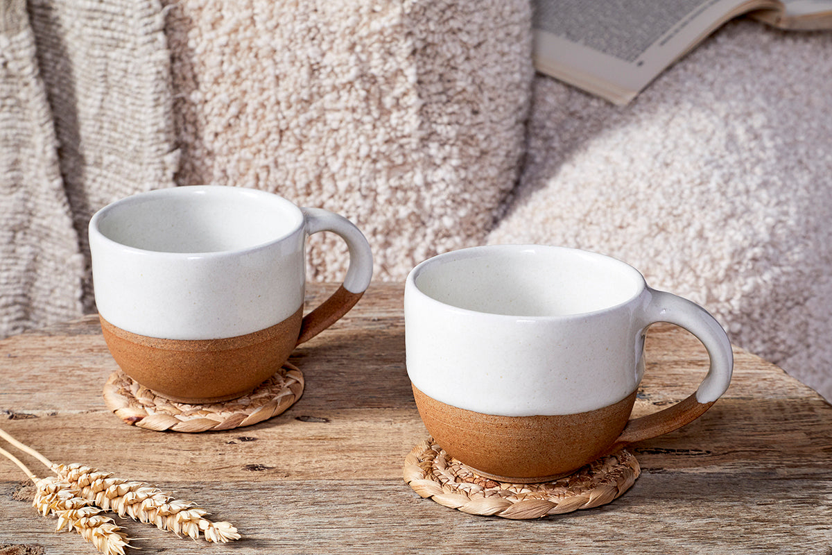 The nkuku Mali Coffee Mug - White (Set of 2) sits on woven coasters atop a rustic wooden table, with cozy beige blankets, an open book, and wheat stalks nearby for a warm, inviting setting.