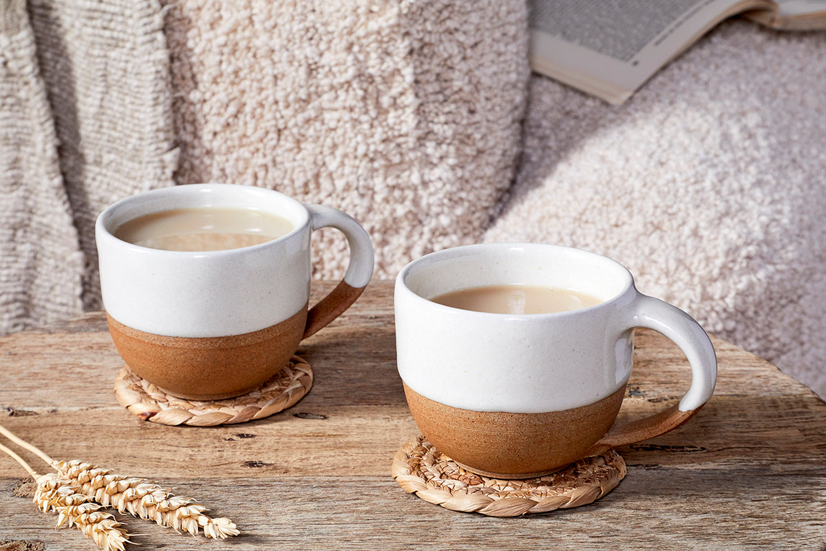 The nkuku Mali Coffee Mug - White (Set of 2) sits on round woven coasters atop a rustic wooden table, with soft beige blankets, an open book, and dried wheat stems adding to the cozy, inviting scene.