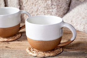 Two Mali White and Terracotta ceramic mugs by nkuku, featuring off-white glaze tops and terracotta bottoms, sit on woven coasters on a wooden table with a fuzzy blanket in the background.