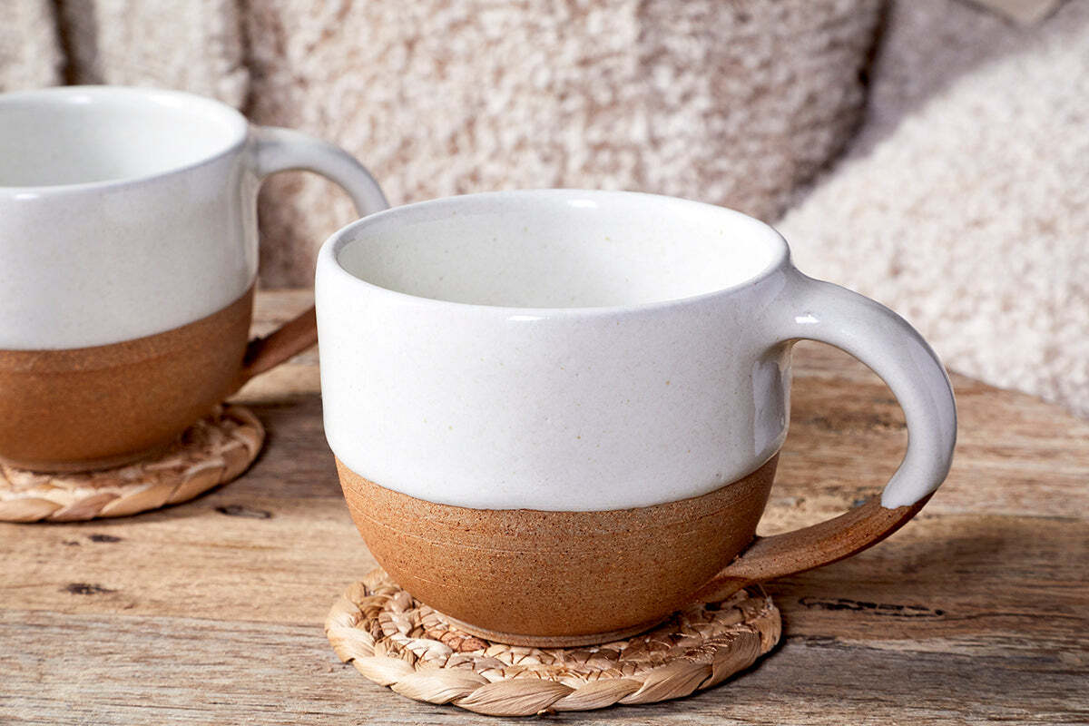 Two Mali White and Terracotta ceramic mugs by nkuku, featuring off-white glaze tops and terracotta bottoms, sit on woven coasters on a wooden table with a fuzzy blanket in the background.