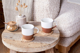 A rustic wooden table displays two nkuku Mali Large Mugs - White, each with white tops and brown bases, placed on woven coasters alongside a small jar of brown sugar and a vase of dried flowers. A cozy textured beige sofa sits in the background.