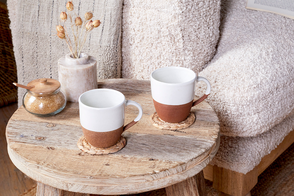 A rustic wooden table displays two nkuku Mali Large Mugs - White, each with white tops and brown bases, placed on woven coasters alongside a small jar of brown sugar and a vase of dried flowers. A cozy textured beige sofa sits in the background.