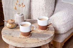 Two nkuku Mali Large Mugs in white sit on coasters atop a round wooden table, beside a brown sugar jar with a wooden lid and a vase of dried flowers. A soft beige armchair and textured blanket appear in the background.