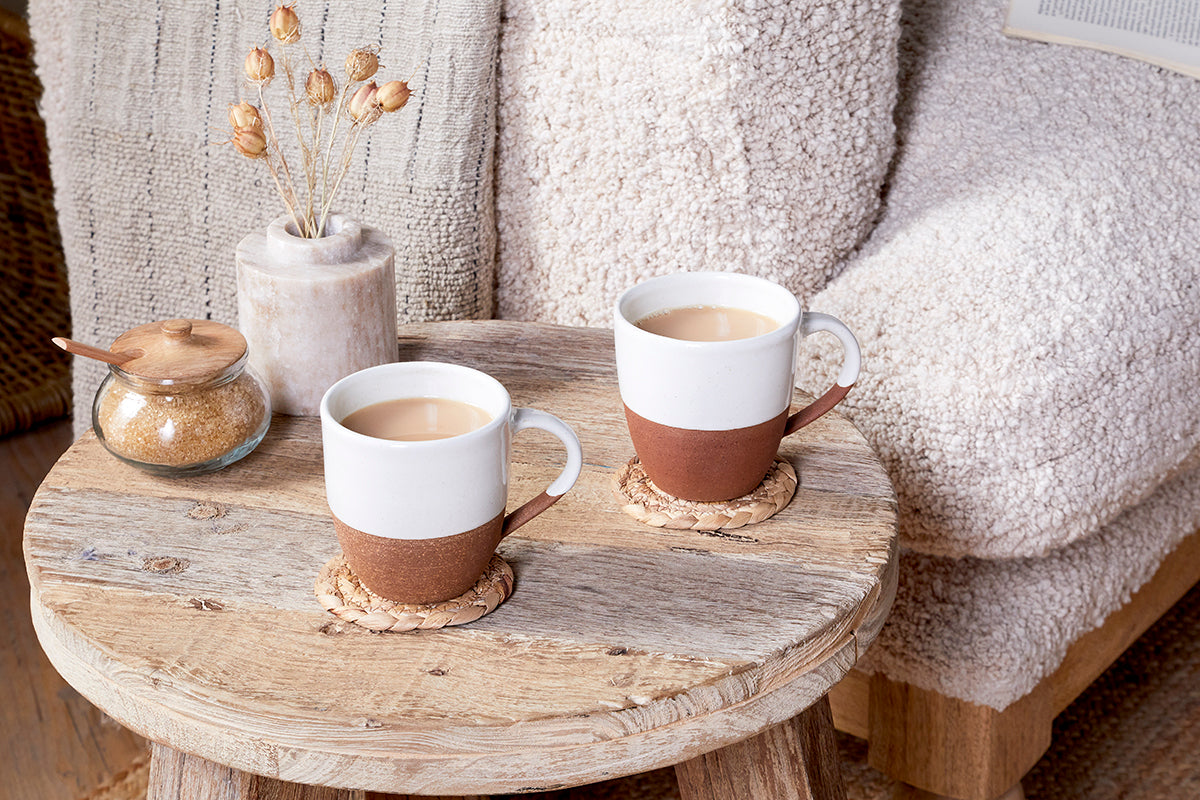 Two nkuku Mali Large Mugs in white sit on coasters atop a round wooden table, beside a brown sugar jar with a wooden lid and a vase of dried flowers. A soft beige armchair and textured blanket appear in the background.