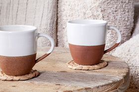 The Mali Large Mug - White (Set of 2) by nkuku, featuring sleek white ceramic design, rests on woven coasters atop a rustic wooden table with soft beige and cream textiles in the background for a cozy ambiance.
