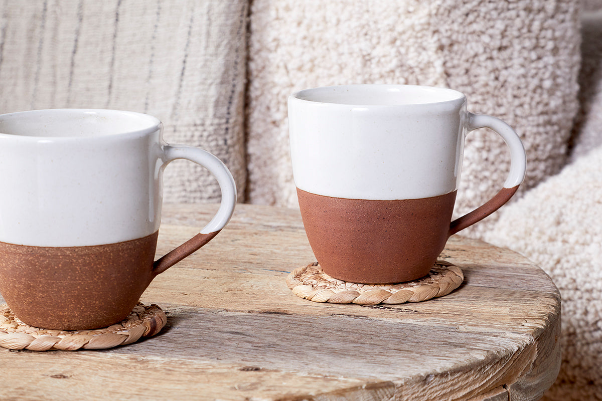 The Mali Large Mug - White (Set of 2) by nkuku, featuring sleek white ceramic design, rests on woven coasters atop a rustic wooden table with soft beige and cream textiles in the background for a cozy ambiance.