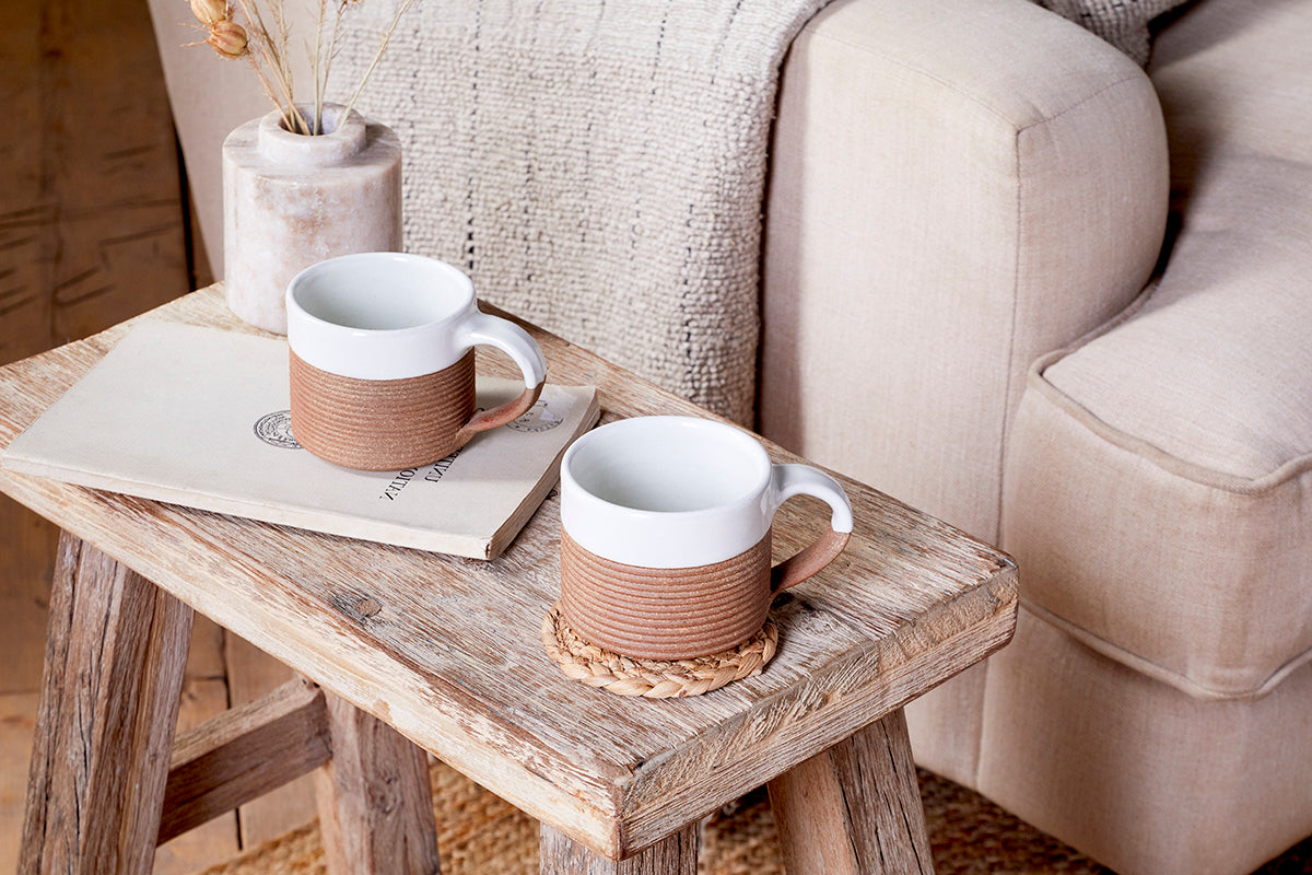 Two Mali Ribbed Coffee Mugs - White by nkuku rest on a rustic wooden stool—one on a woven coaster, beside a closed book. A neutral couch and dried flowers add to the cozy, natural vibe.