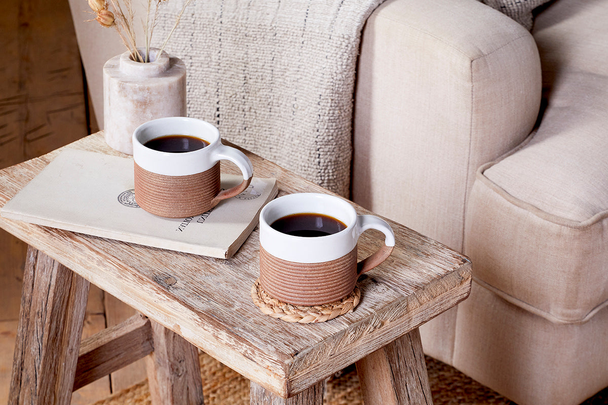 A set of two nkuku Mali Ribbed Coffee Mugs in white sit on a rustic wooden table—one on a coaster, one on a closed book—with a beige couch and dried flowers completing the cozy, inviting scene.