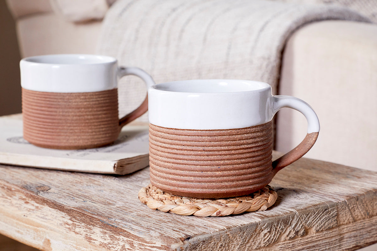 Two nkuku Mali Ribbed Coffee Mugs in white sit on a rustic wooden table—one on a woven coaster, the other on a closed book—with a beige knitted blanket draped over a sofa in the softly blurred background.