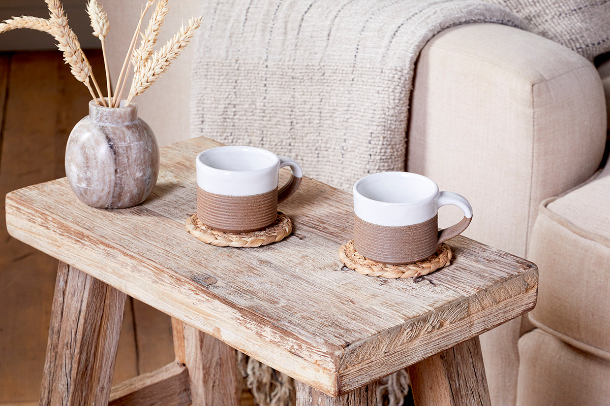 A rustic wooden coffee table displays nkuku's Mali Ribbed Espresso Mugs - White (Set of 2) on woven coasters, beside a ceramic vase with dried wheat. A beige sofa and textured blanket create a warm, inviting setting.