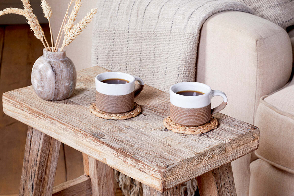 A rustic wooden table holds two nkuku Mali Ribbed Espresso Mugs in white on woven coasters beside a beige sofa. A textured gray blanket drapes over the arm, and a small vase with dried wheat adds cozy warmth.