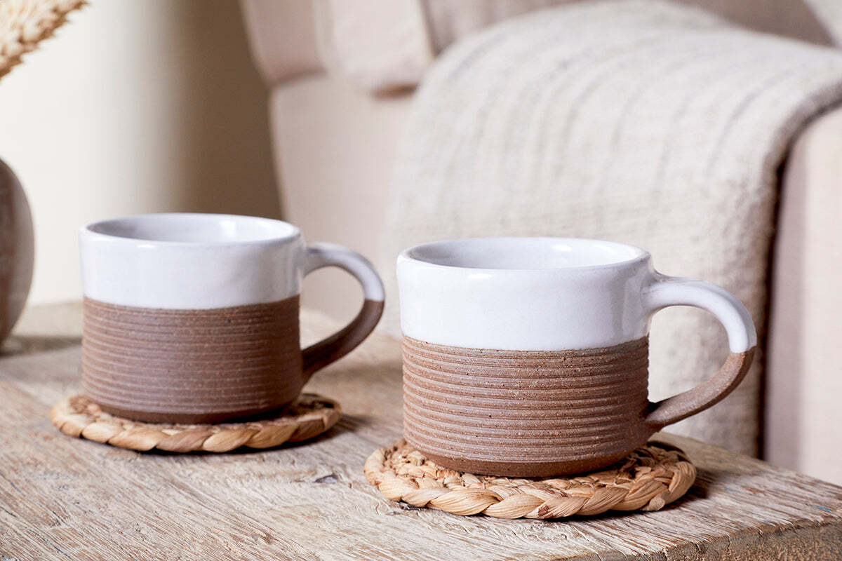 Two nkuku Mali Ribbed Espresso Mugs - White rest on woven coasters atop a wooden table, while a beige chair with a cream blanket in the background enhances the cozy vibe, showcasing the charm of the Mali collection.