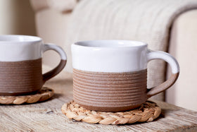 The nkuku Mali Ribbed Espresso Mugs in white (set of 2) rest on woven coasters atop a rustic wooden table, while a soft beige blanket drapes over a sofa in the background, creating a cozy atmosphere.
