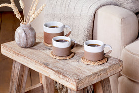 Two nkuku Mali Ribbed Espresso Mugs in white sit on woven coasters atop a rustic wooden table, with coffee poured inside. A small vase with dried wheat and a beige sofa with a blanket complete the cozy living room setting.