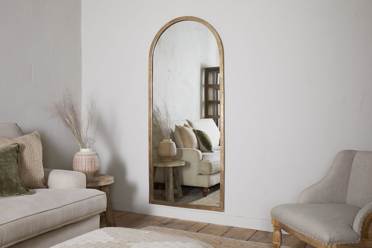 A neutral-toned living room features the nkuku Murwara Full Length Mirror in Antique Brass, reflecting a white sofa with green pillows, a side table, and a vase with dried branches. Wooden floors and light furniture create a calm, cozy atmosphere.