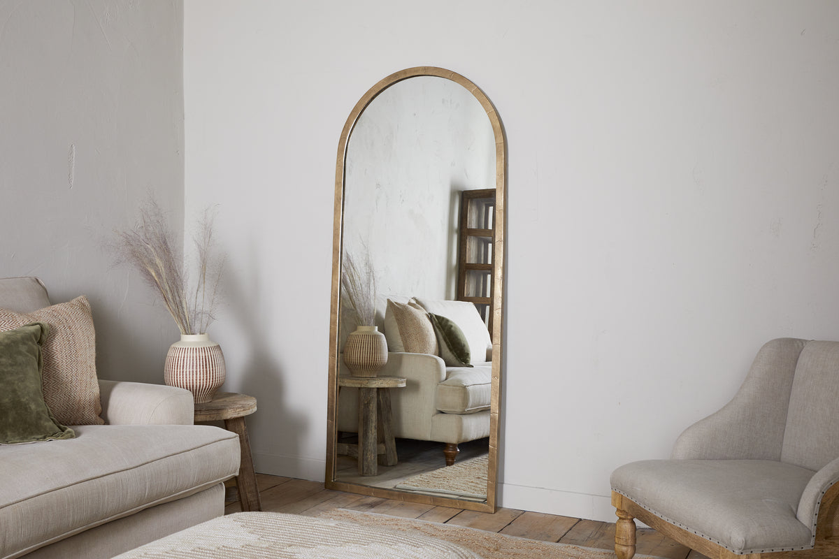The nkuku Murwara Full Length Mirror in Antique Brass leans against a white wall, reflecting a beige sofa, green pillow, and décor. A wicker vase with dried grass sits on a wooden stool beside light wood floors and chairs.