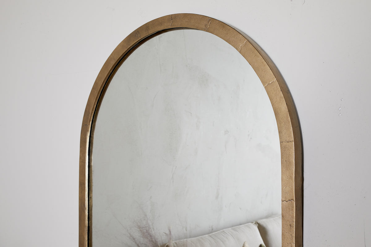 The Murwara Full Length Mirror by nkuku, featuring an arched antique brass frame, hangs on a white wall. Its reflection shows a light sofa and subtle decor, adding minimalist elegance to the space.