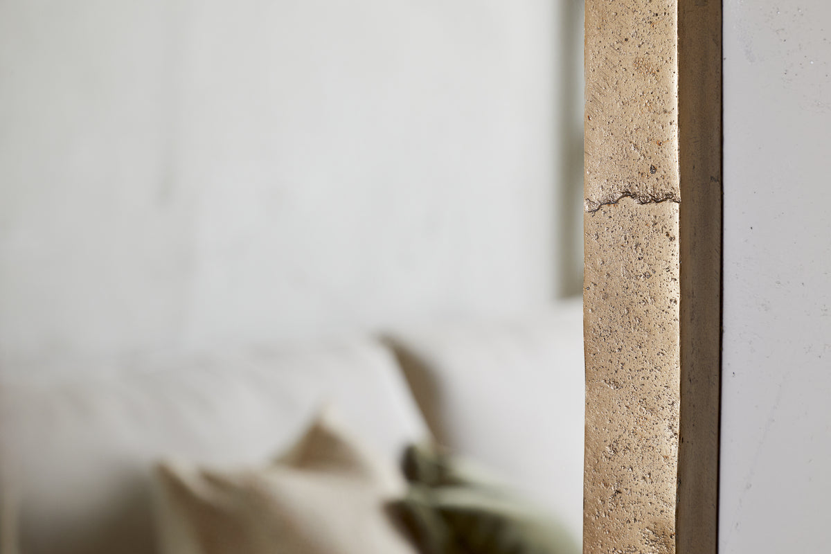 Close-up of a textured beige concrete wall with a visible crack. In the blurred background, the nkuku Murwara Full Length Mirror in Antique Brass stands beside a white sofa and neutral pillows, creating a soft, minimalist interior.