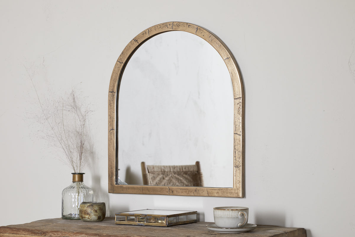 A Murwara Wall Mirror by nkuku in Antique Brass hangs on a white wall above a rustic wooden table topped with a glass vase of dried branches, a decorative box, a candle, and a cup with saucer.