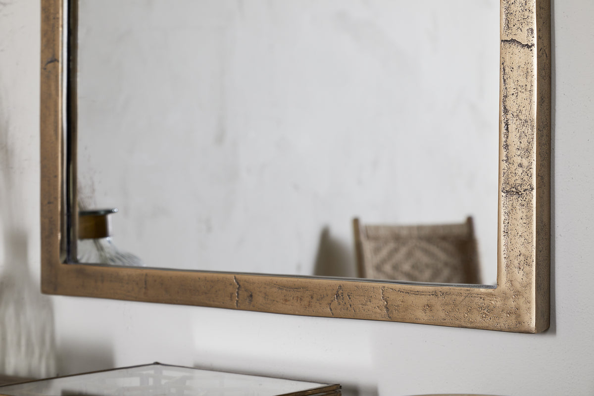 A close-up of the nkuku Murwara Wall Mirror in Antique Brass, mounted on a white wall. Its arched frame reflects part of a patterned chair and glass décor, creating a scene with soft neutral tones.