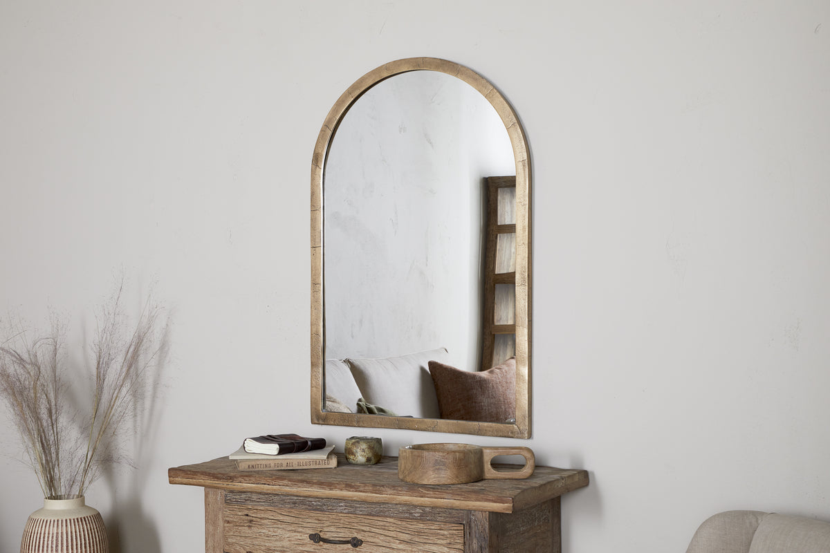 The nkuku Murwara Wall Mirror in antique brass with an arched wooden frame hangs above a rustic table with books, a candle, and a tray. A vase of dried grasses sits to the left, while the cozy sofa is reflected in the mirror.