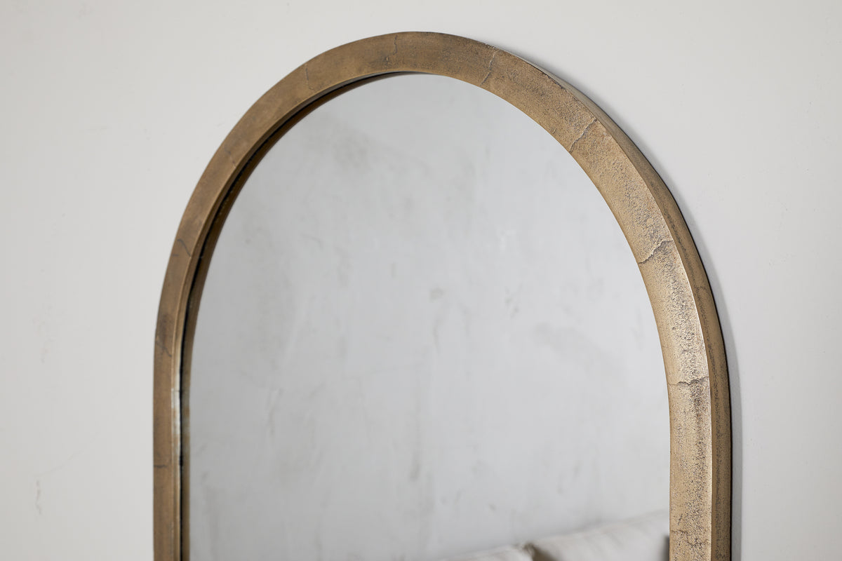 A close-up of the nkuku Murwara Wall Mirror in Antique Brass, reflecting a light-textured wall and the upper edge of a pale sofa.