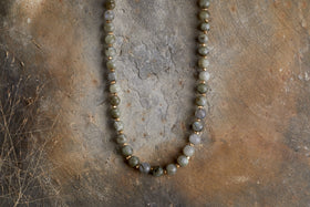 The Marai Necklace - Labradortite by nkuku features round, earthy labradorite beads arranged in a semicircle, displayed on a rustic brown textured surface for an elegant look.