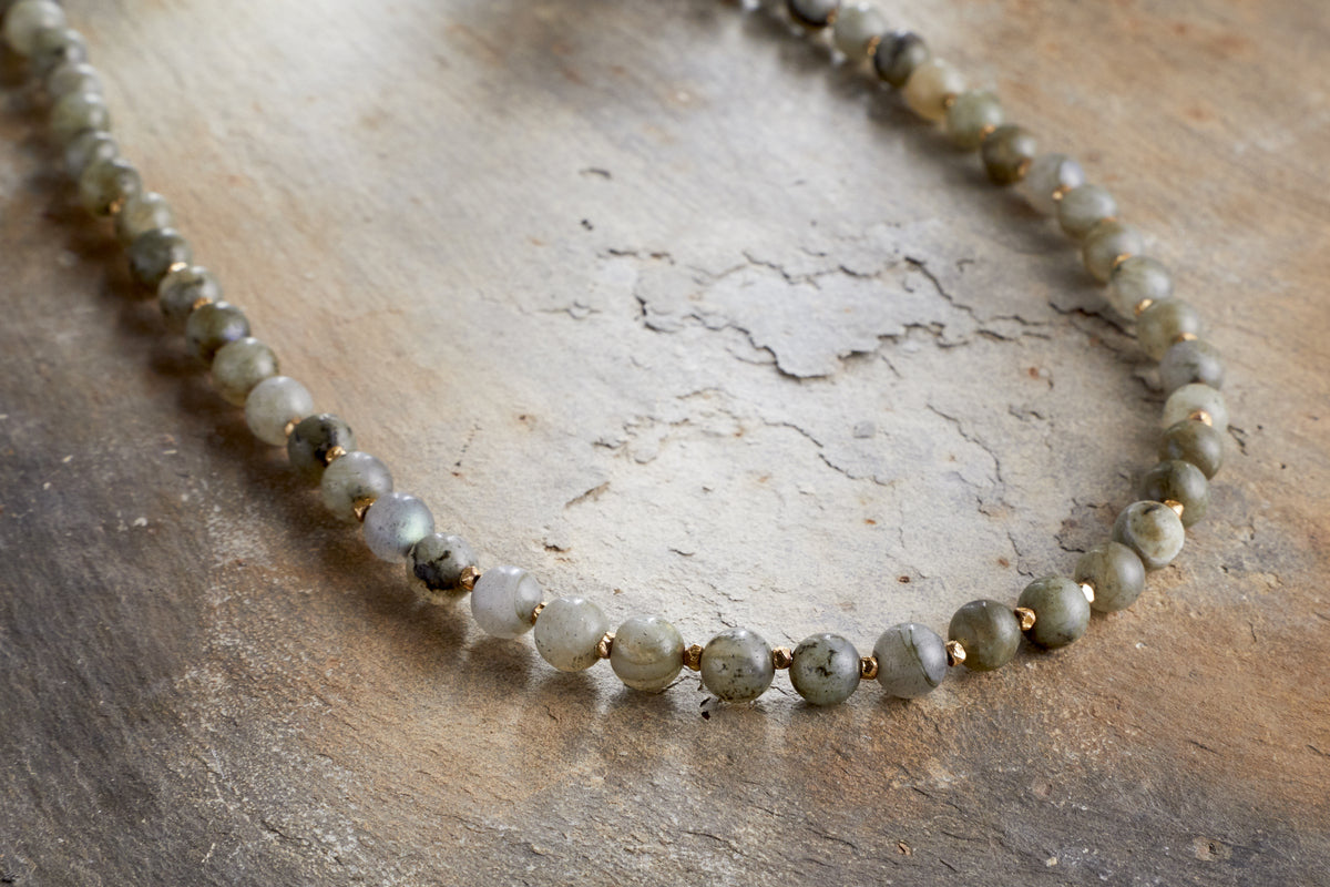 The Marai Necklace - Labradorite by nkuku showcases round, translucent grey-green labradorite stones and small gold spacer beads, elegantly arranged on a textured surface—an exquisite piece that embodies natural beauty.