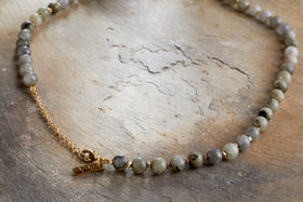 The Marai Necklace - Labradorite by nkuku features round, speckled grey labradorite beads, a short gold chain extension, and a small rectangular gold tag—an elegant piece displayed on a textured stone-like surface.