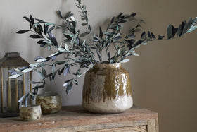 The Melina Reactive Glaze Vase in Olive & Natural by nkuku holds green leafy branches on a rustic wooden surface, accompanied by two small ceramic cups and a lantern for a cozy, natural decor scene.