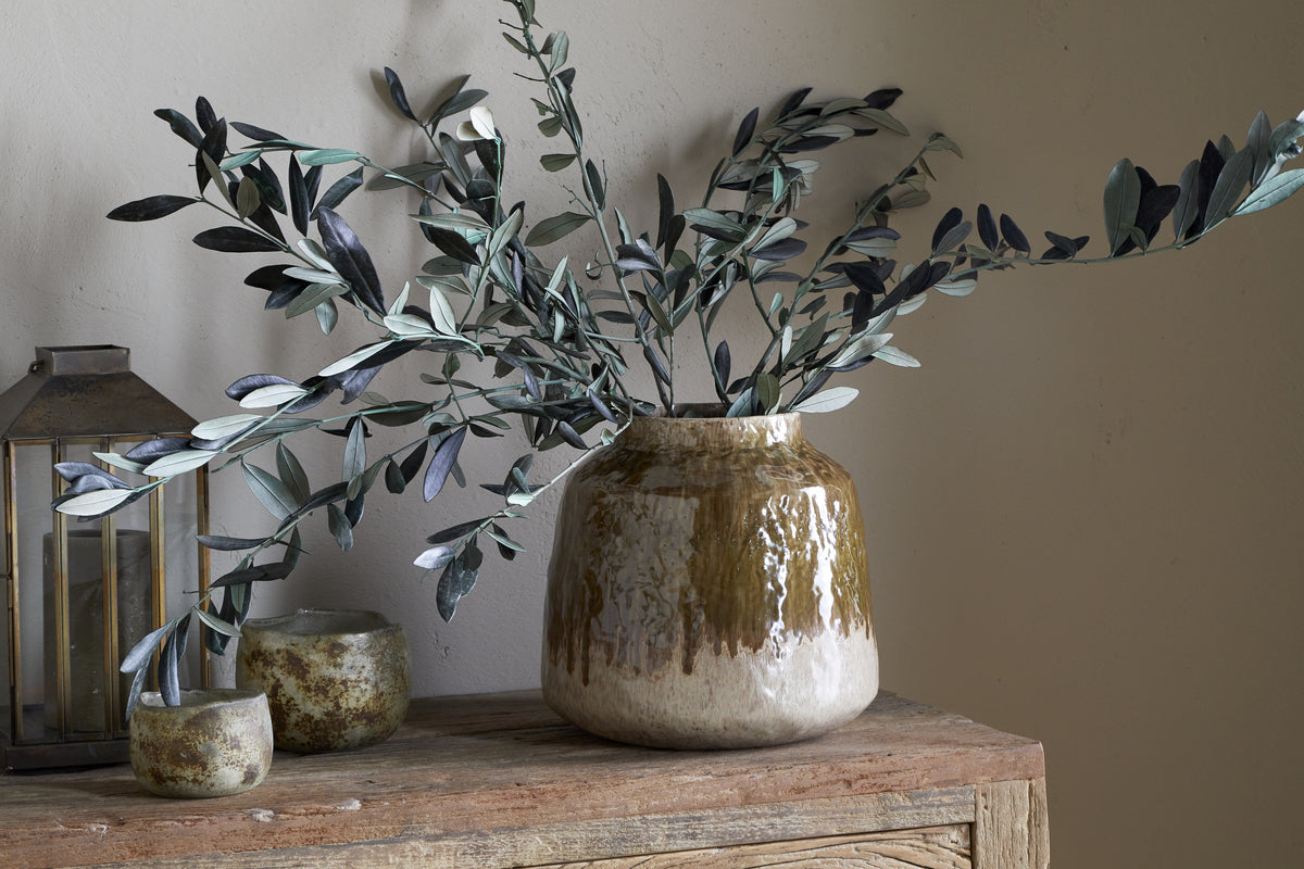 The Melina Reactive Glaze Vase in Olive & Natural by nkuku holds green leafy branches on a rustic wooden surface, accompanied by two small ceramic cups and a lantern for a cozy, natural decor scene.