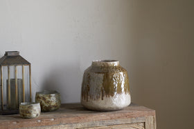 The Melina Reactive Glaze Vase in Olive & Natural by nkuku sits on a rustic wooden surface beside two small textured cups and a metal lantern, set against a plain light-colored wall.