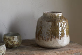 The nkuku Melina Reactive Glaze Vase in Olive & Natural sits on a rustic wooden surface beside two small textured ceramic cups, set against a plain light-colored wall.