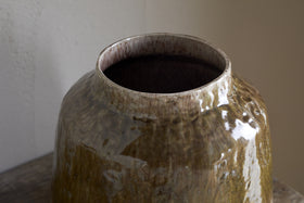 The Melina Reactive Glaze Vase by nkuku, in Olive & Natural, features a large, glossy finish with an uneven surface and wide opening. This decorative vase sits empty on a wooden table against a light wall.