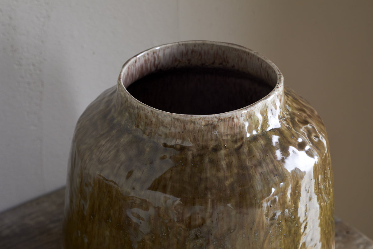 The Melina Reactive Glaze Vase by nkuku, in Olive & Natural, features a large, glossy finish with an uneven surface and wide opening. This decorative vase sits empty on a wooden table against a light wall.