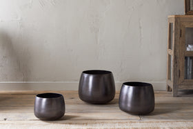 Three nkuku Maliki Plant Pots in aged bronze with a matte finish, each a different height, are arranged on a wooden floor by a light beige wall. A rustic shelf with books is visible to the right, complementing the minimalist look.