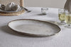 The nkuku Malia Large Oval Platter in cream, featuring an organic shape and textured rim, rests on a light gray tablecloth beside two pale drinks, a lit candle, and a place setting with plate, napkin, and woven placemat.