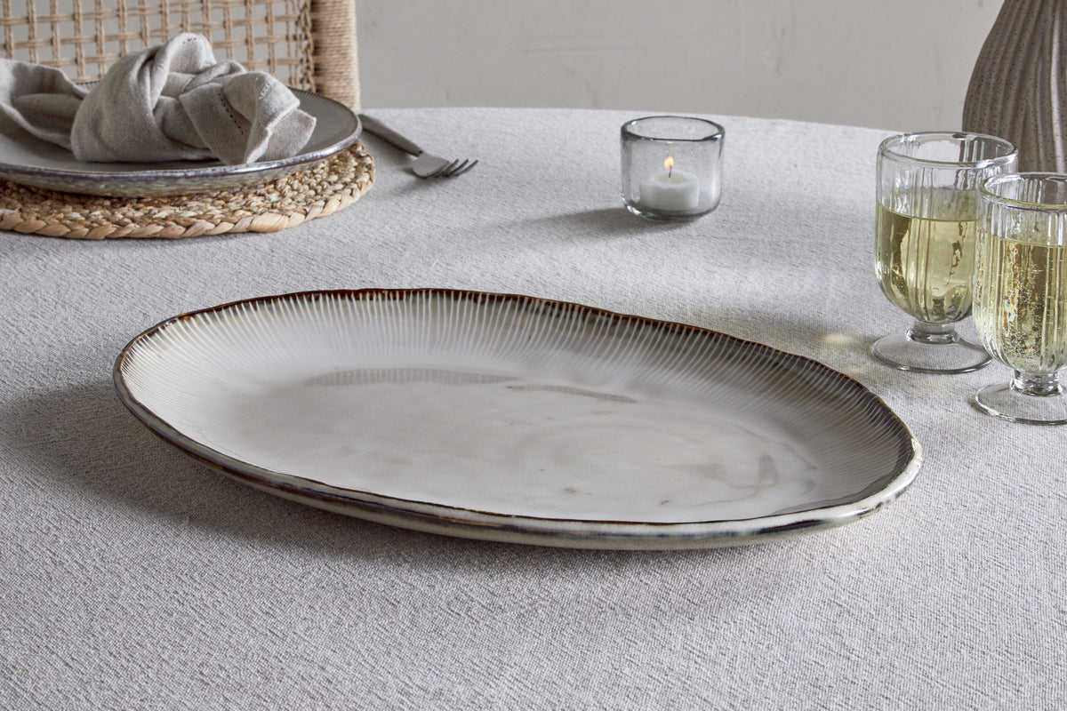 The nkuku Malia Large Oval Platter in cream, featuring an organic shape and textured rim, rests on a light gray tablecloth beside two pale drinks, a lit candle, and a place setting with plate, napkin, and woven placemat.