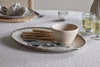 The nkuku Malia Large Oval Platter in Cream displays breadsticks, a wooden bowl, and greenery on a textured tablecloth. Around it are two sparkling drinks in glasses, a small candle, and a plate with a napkin.